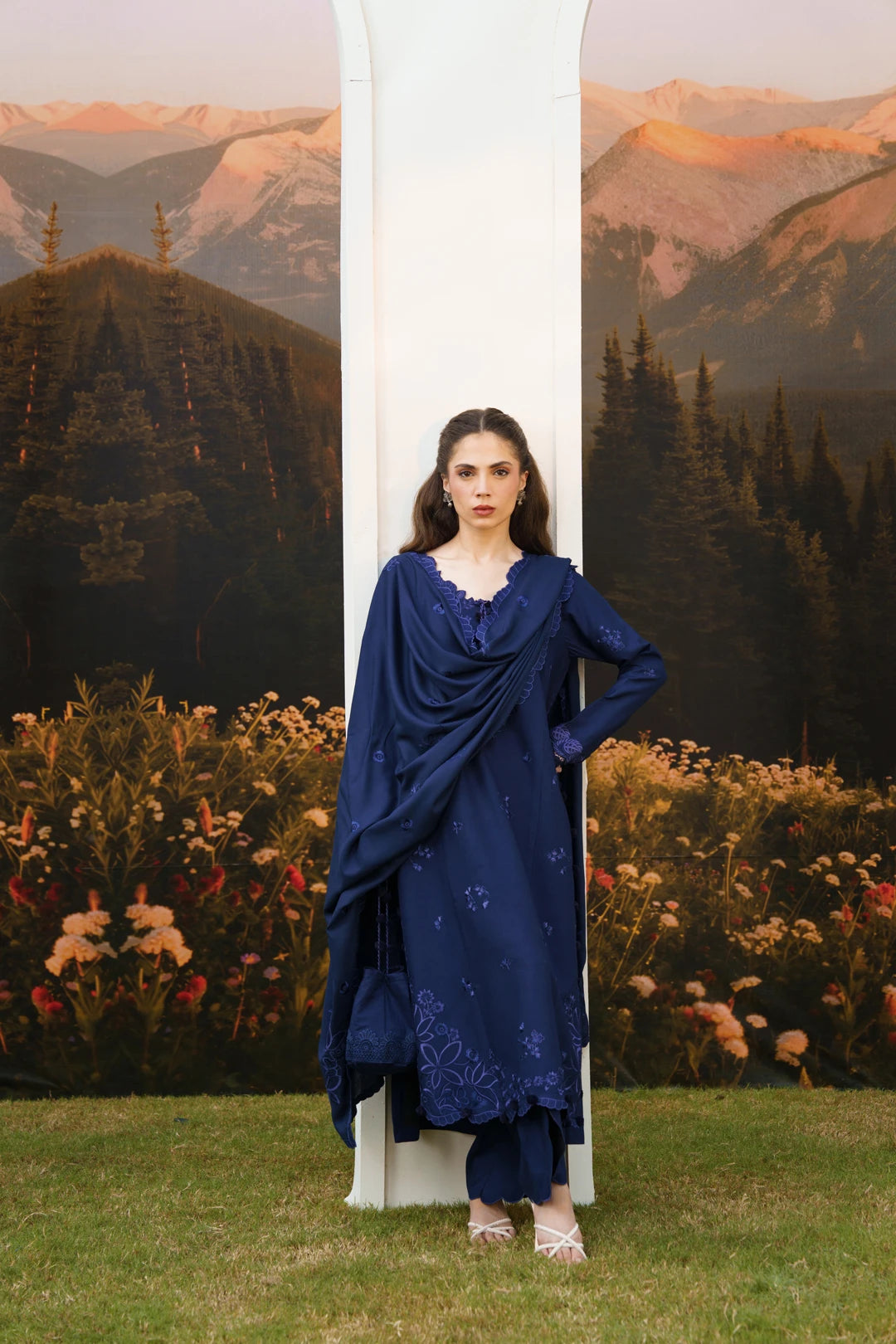 A woman stands outdoors in front of a scenic mountain backdrop and flowerbed, wearing a deep navy blue traditional outfit with a draped shawl and matching pants. A large white archway is behind her.