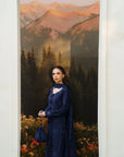 A woman stands in front of a large scenic backdrop showing mountains, trees, and flowers, wearing a long navy blue embroidered dress with a matching headscarf and holding a matching handbag.