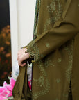 Close-up of a person wearing an olive green embroidered traditional outfit with matching belt and holding a white bag, set against a softly blurred background with pink flowers.