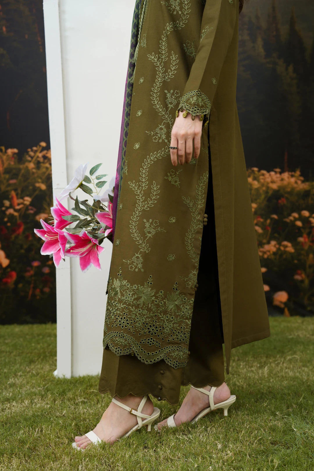 A person outdoors wearing an olive green traditional outfit with matching embroidered kurta and trousers, paired with white heeled sandals. The background shows grass, flowers, and a dark forest mural.