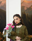 A woman models an olive green embroidered kurta with a matching purple dupatta, holding a pink flower bouquet in front of a scenic mountain backdrop.