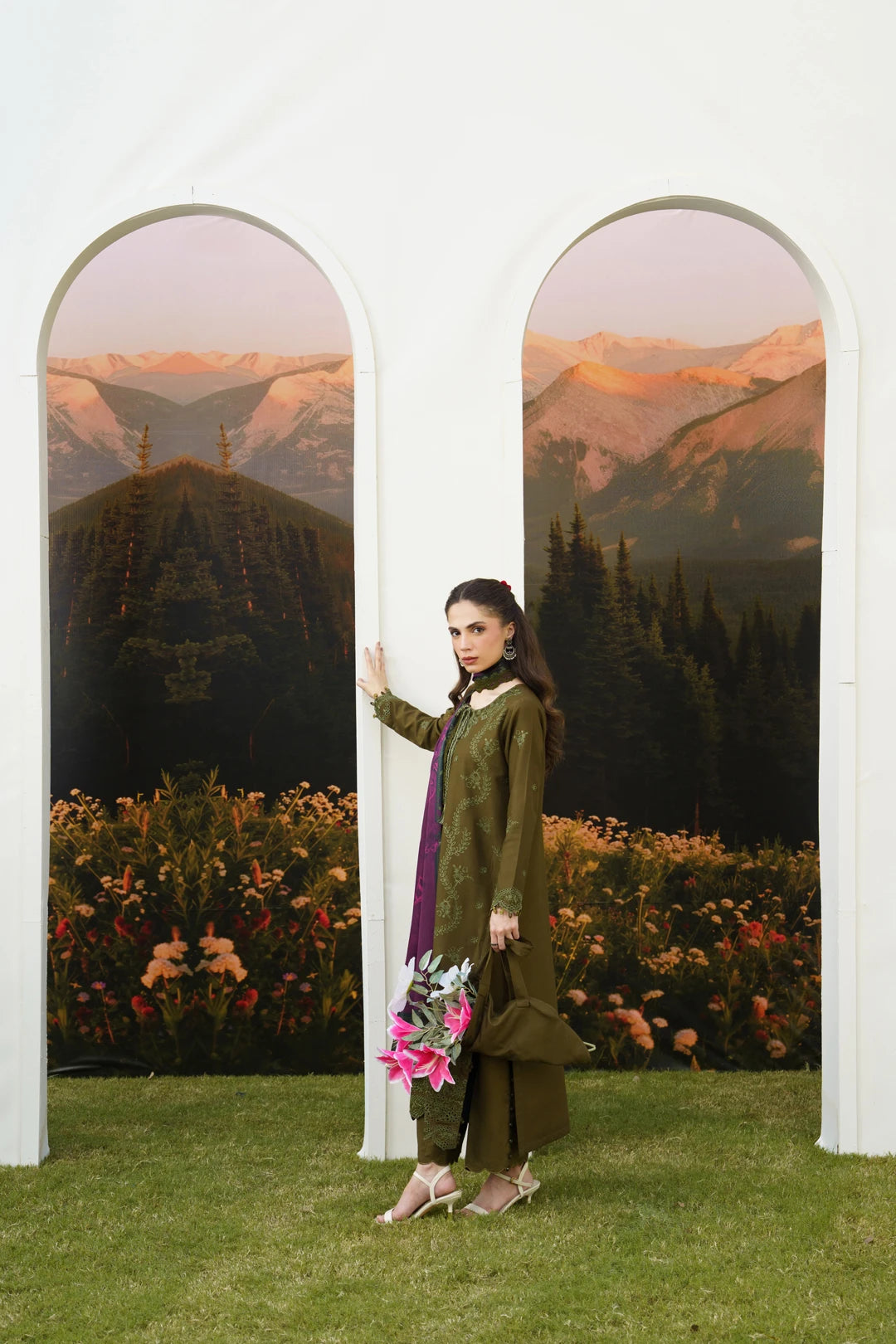 A woman models an olive green traditional outfit with a purple dupatta, standing in front of a white architectural backdrop featuring two arched panels with mountain scenery.