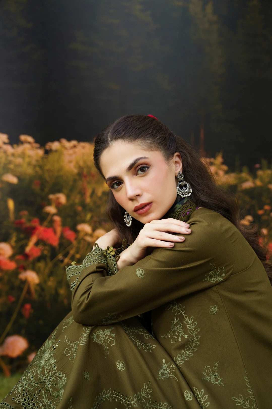 A woman wearing an olive green embroidered traditional outfit with matching embroidered sleeves, posed in front of a blurred outdoor garden background with flowers and trees.