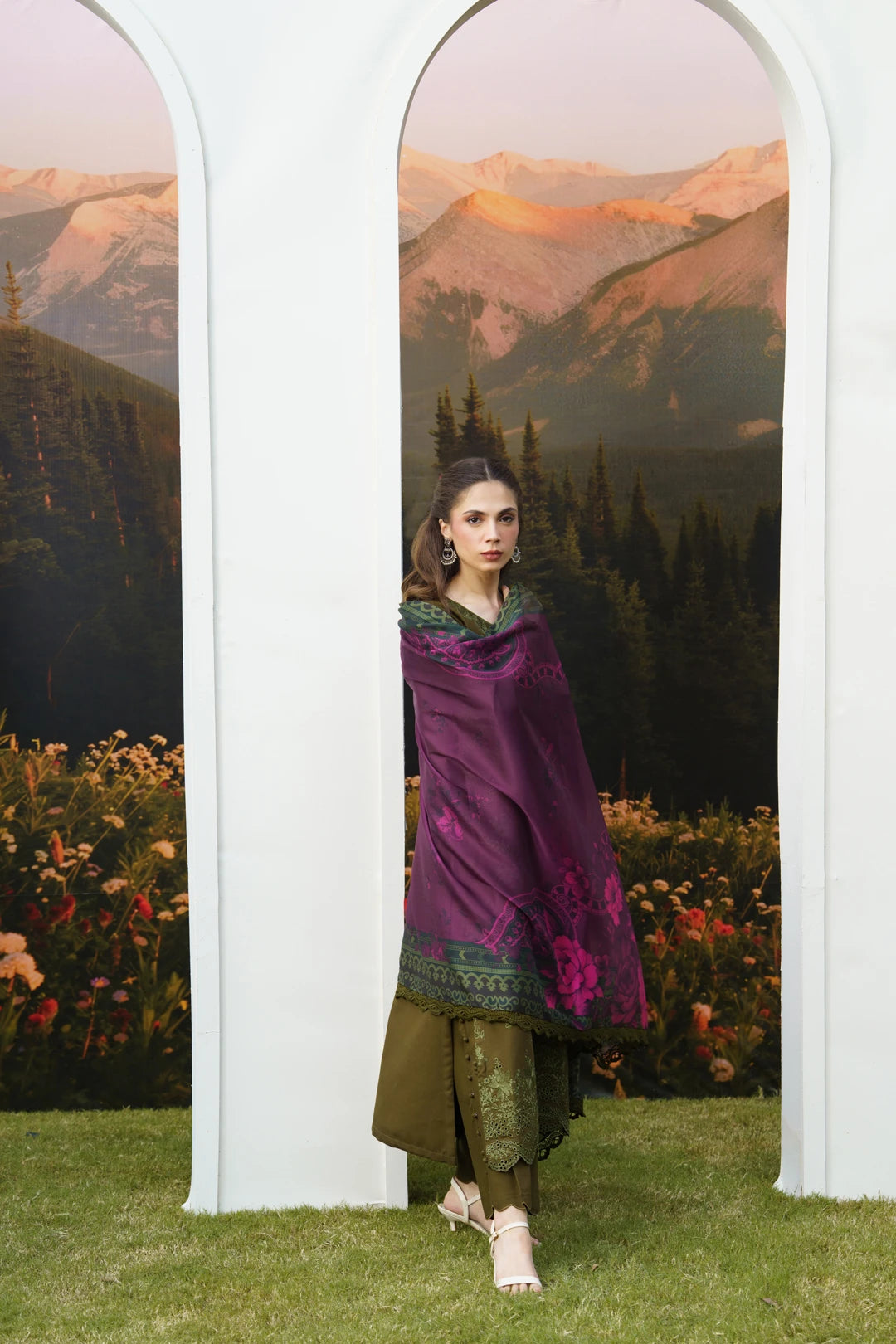 A woman models a traditional South Asian outfit consisting of a deep purple embroidered dupatta, olive green trousers, and a green patterned top, standing between two white arched frames with a mountain landscape backdrop.