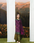 A woman models a traditional South Asian outfit consisting of a deep purple embroidered dupatta, olive green trousers, and a green patterned top, standing between two white arched frames with a mountain landscape backdrop.