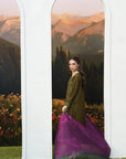 A woman models a traditional South Asian outfit consisting of an olive green kurta and a purple printed dupatta, standing between two white arched frames with a scenic mountain backdrop.