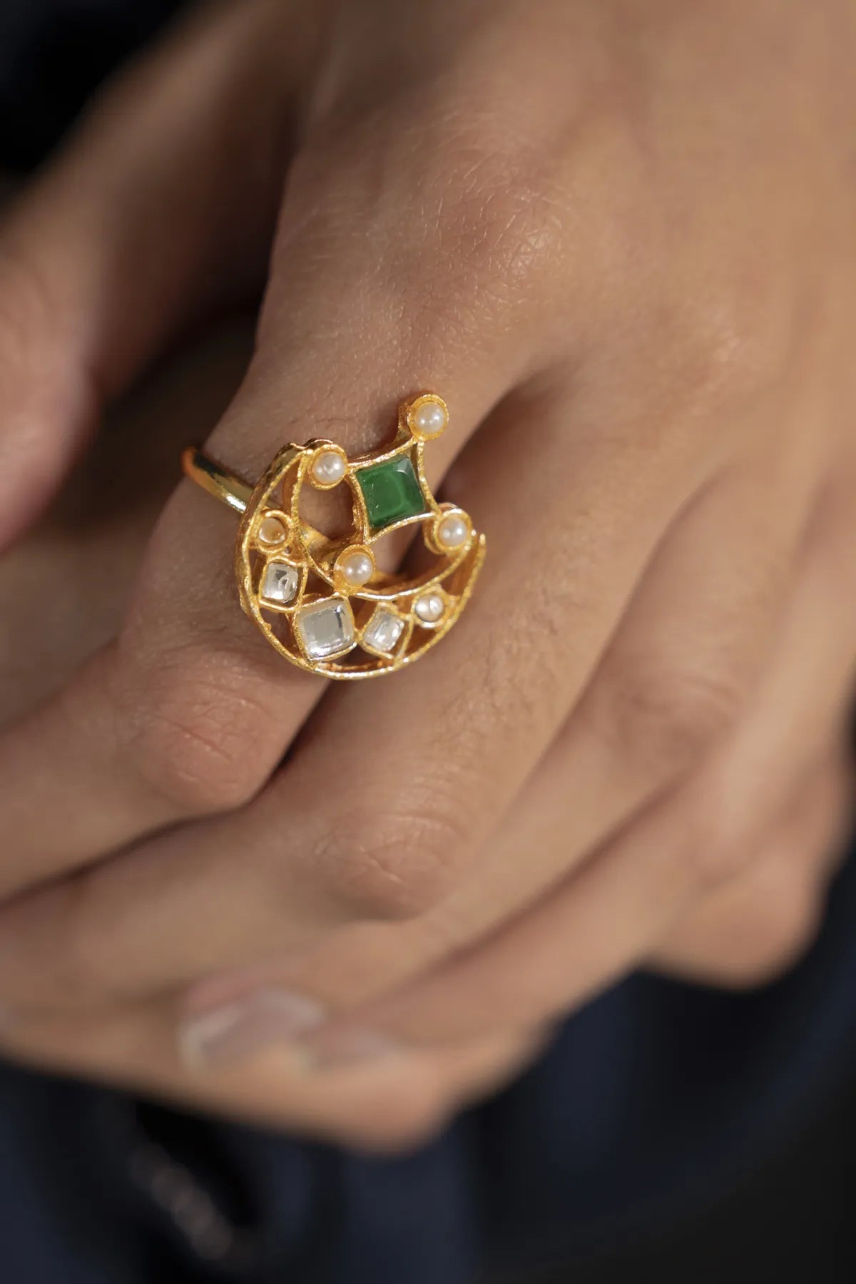 Gold-tone crescent kundan ring with green center stone and pearl accents, close-up on hand.