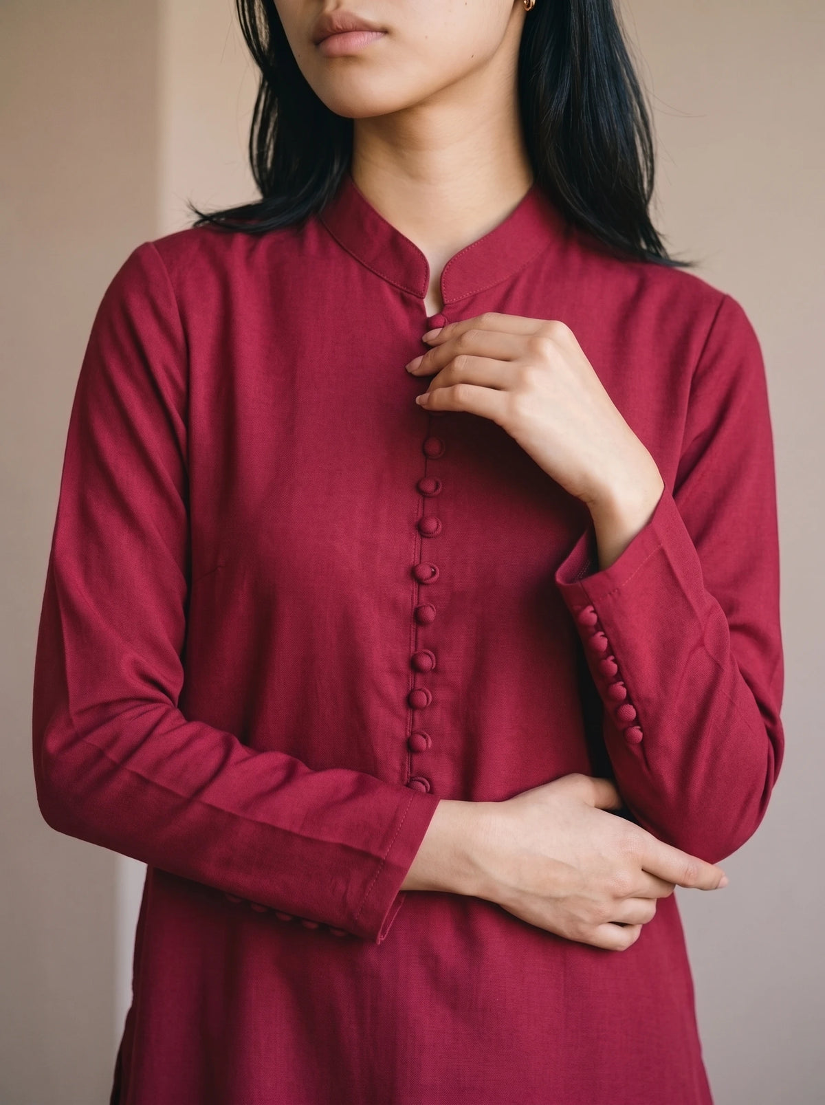 A woman wearing a long-sleeved, deep red button-up tunic with a mandarin collar, shown from the chest down against a neutral beige background.