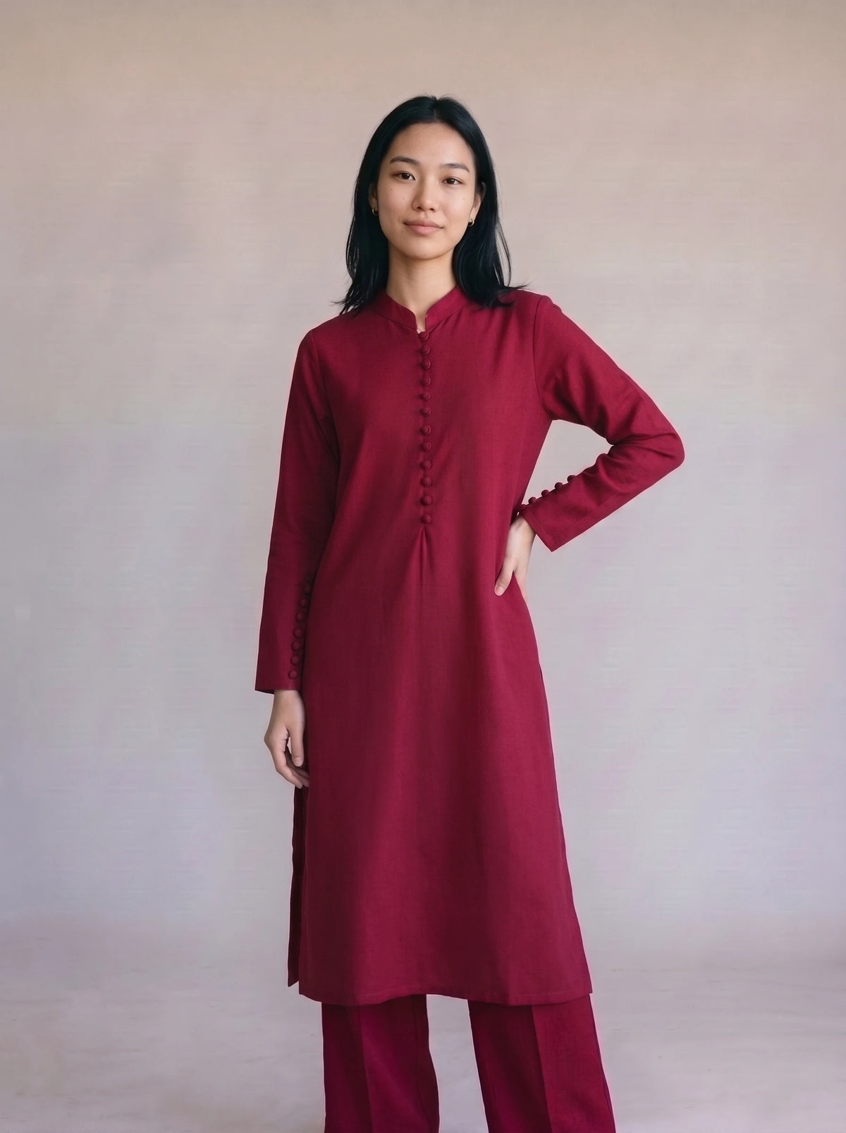 A woman modeling a long-sleeved deep red kurta with a buttoned mandarin collar and matching red trousers, standing against a neutral beige studio backdrop.