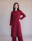 A woman modeling a long-sleeved deep red kurta with a buttoned mandarin collar and matching red trousers, standing against a neutral beige studio backdrop.
