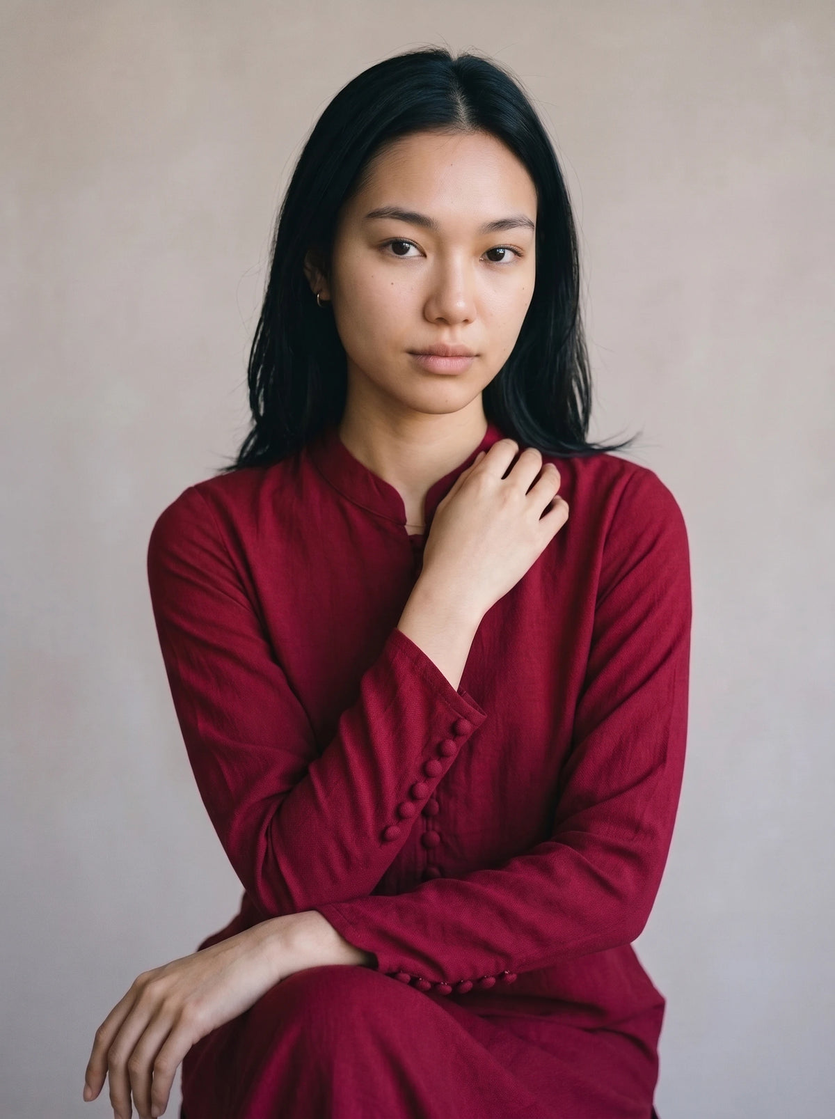 A woman with long black hair wearing a dark red long-sleeve top with decorative buttons on the cuffs, posing against a neutral beige background.