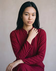 A woman with long black hair wearing a dark red long-sleeve top with decorative buttons on the cuffs, posing against a neutral beige background.