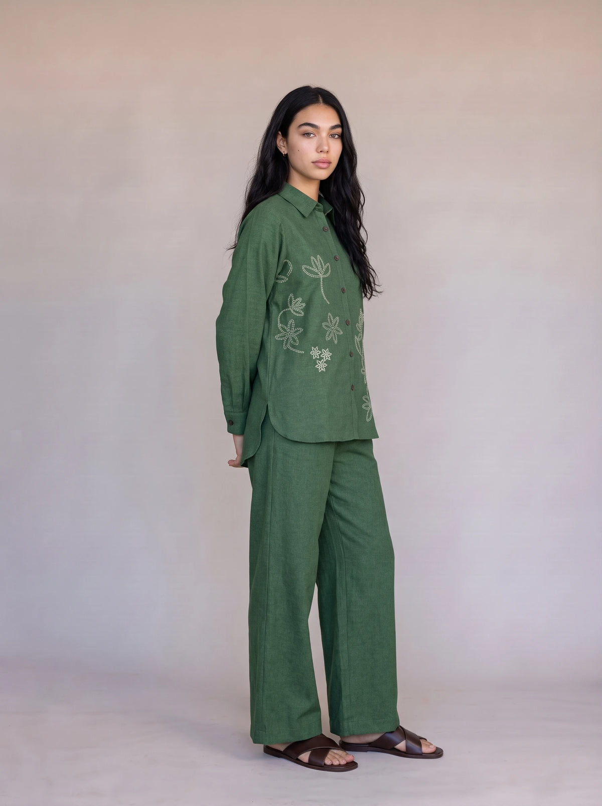 A woman modeling a matching green two-piece set consisting of a long-sleeve embroidered top and wide-leg trousers, standing against a neutral beige studio backdrop.