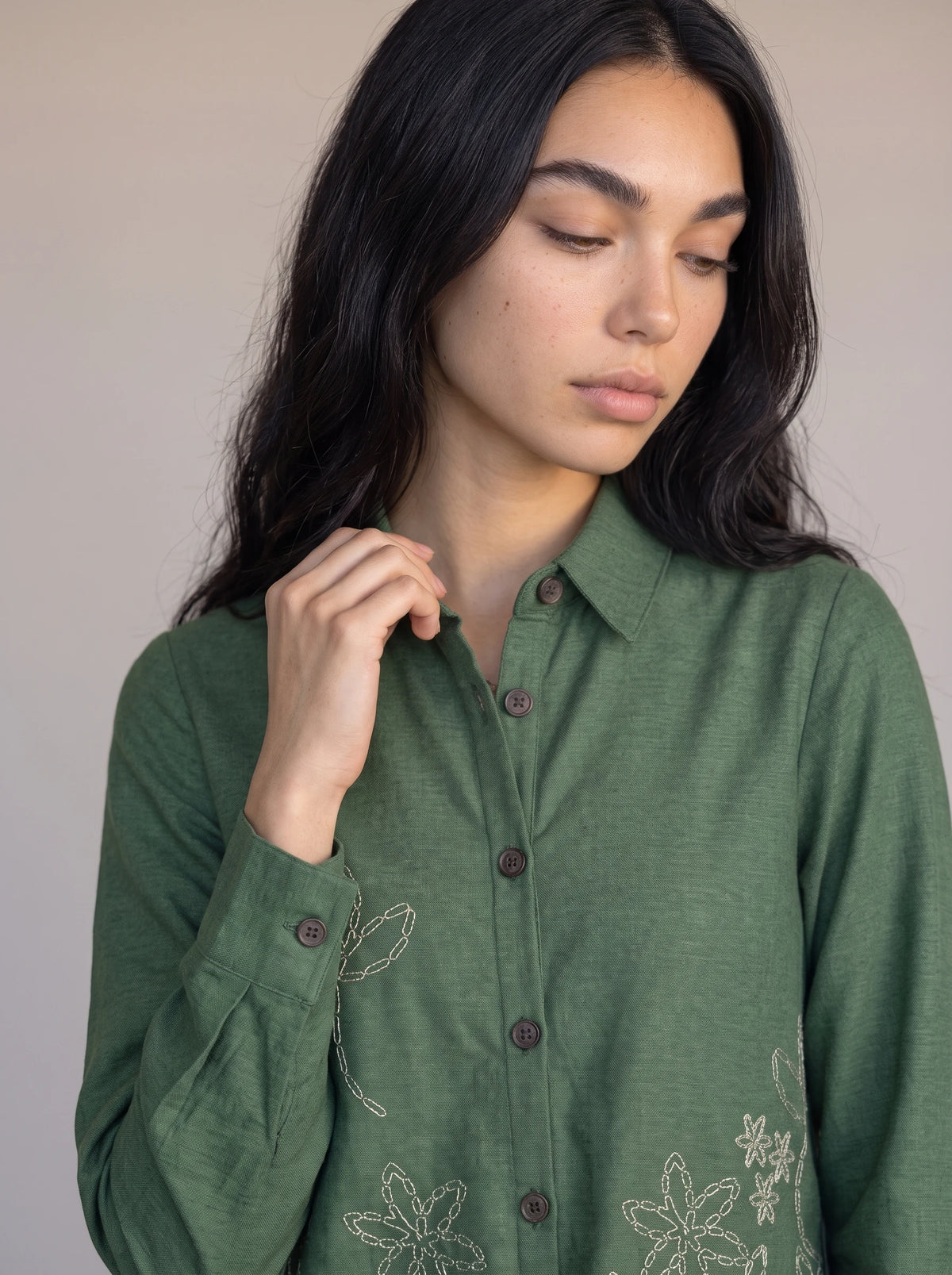 A woman wearing a sage green long-sleeve button-up shirt with white floral embroidery on the front, posed against a neutral beige background.