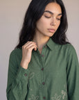 A woman wearing a sage green long-sleeve button-up shirt with white floral embroidery on the front, posed against a neutral beige background.