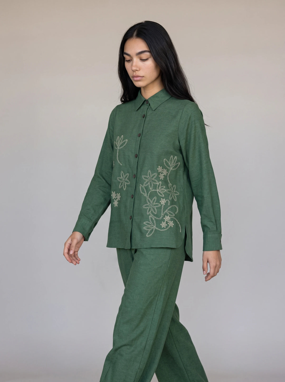A woman modeling a matching green two-piece set consisting of a long-sleeve button-up shirt with white floral embroidery and matching wide-leg trousers, set against a plain light gray studio background.