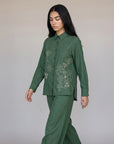 A woman modeling a matching green two-piece set consisting of a long-sleeve button-up shirt with white floral embroidery and matching wide-leg trousers, set against a plain light gray studio background.