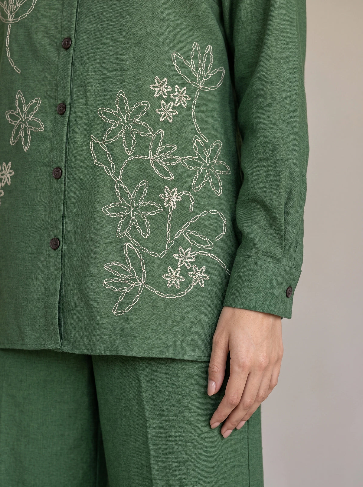 A close-up of a person wearing a green long-sleeve button-front top with white floral embroidery and matching green pants, against a plain light gray background.