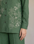 A close-up of a person wearing a green long-sleeve button-front top with white floral embroidery and matching green pants, against a plain light gray background.