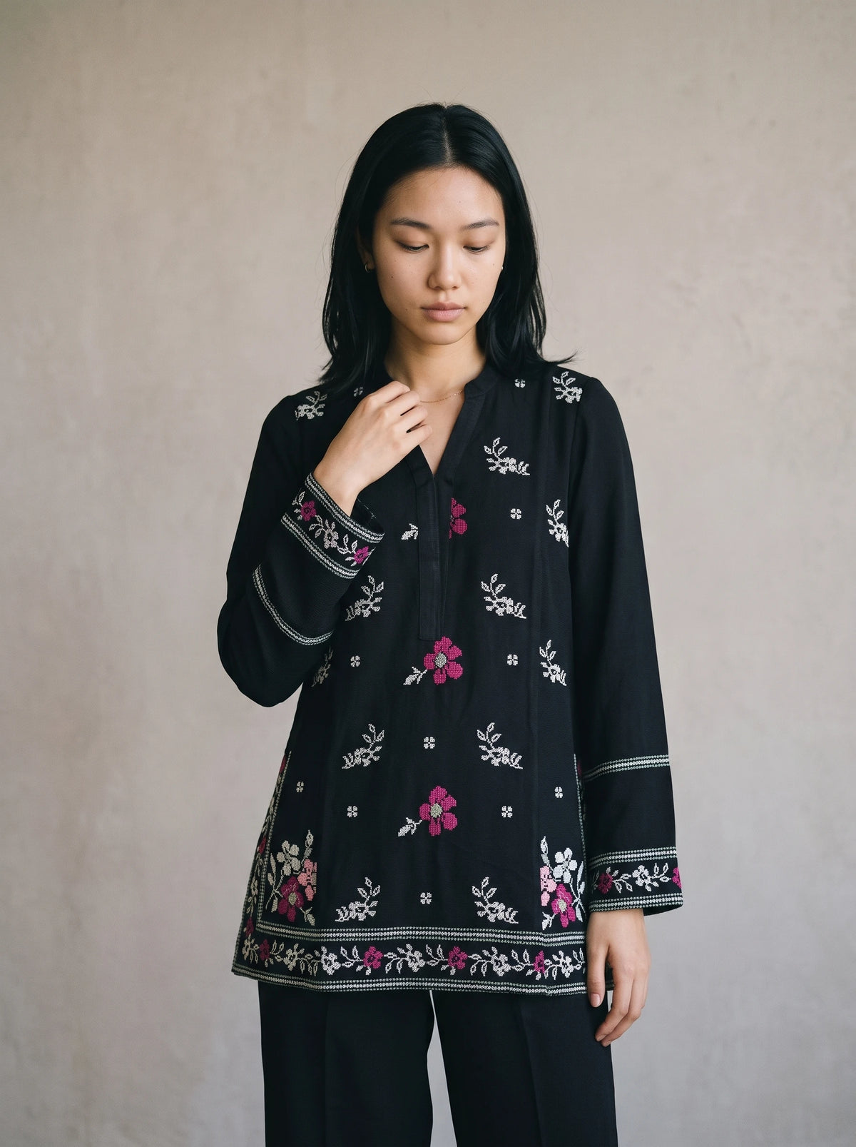 A woman wearing a black long-sleeve tunic top with pink and white floral embroidery, paired with black pants, standing against a neutral textured beige wall.