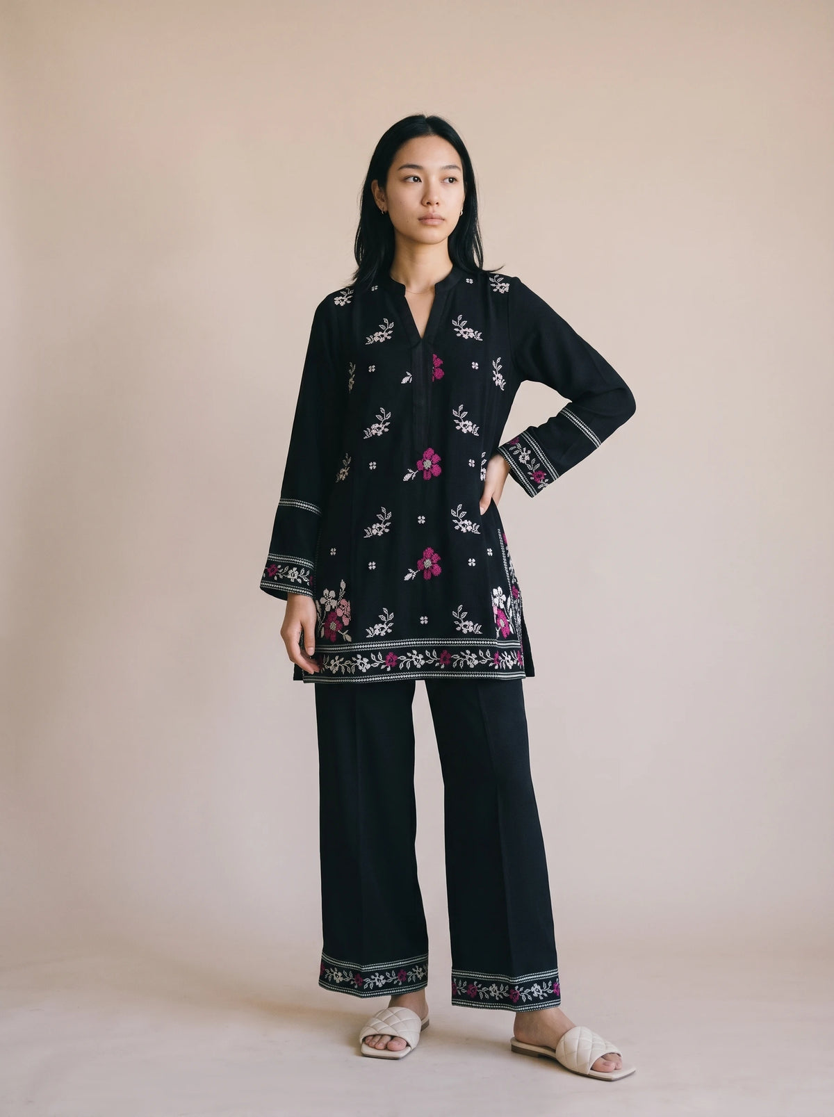 A woman modeling a black two-piece kurta and trouser set with colorful floral embroidery and patterned borders, standing against a solid beige studio backdrop.