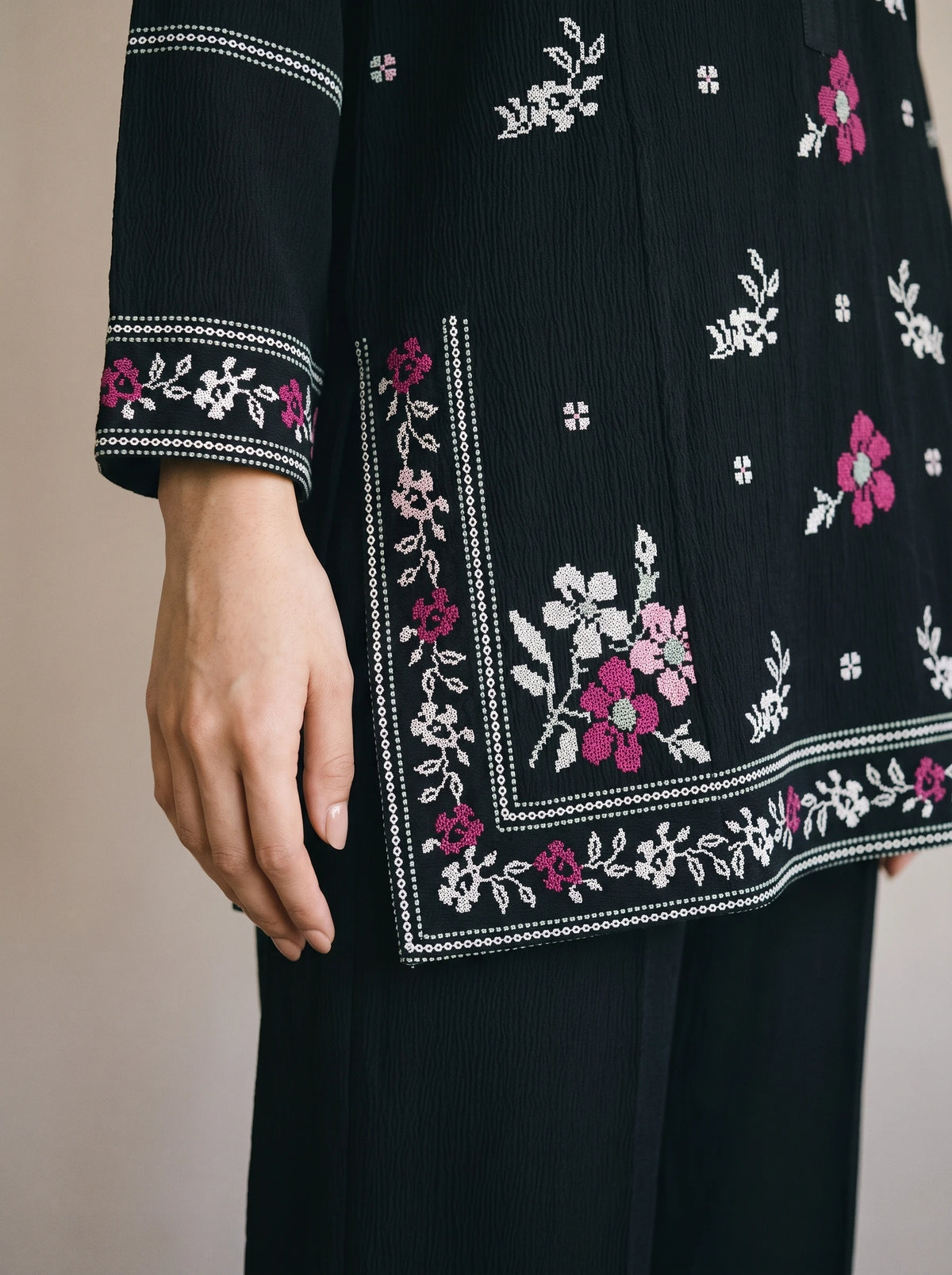 Close-up of a black embroidered tunic or kurta with pink and white floral patterns, shown on a model with a matching black skirt against a plain beige background.