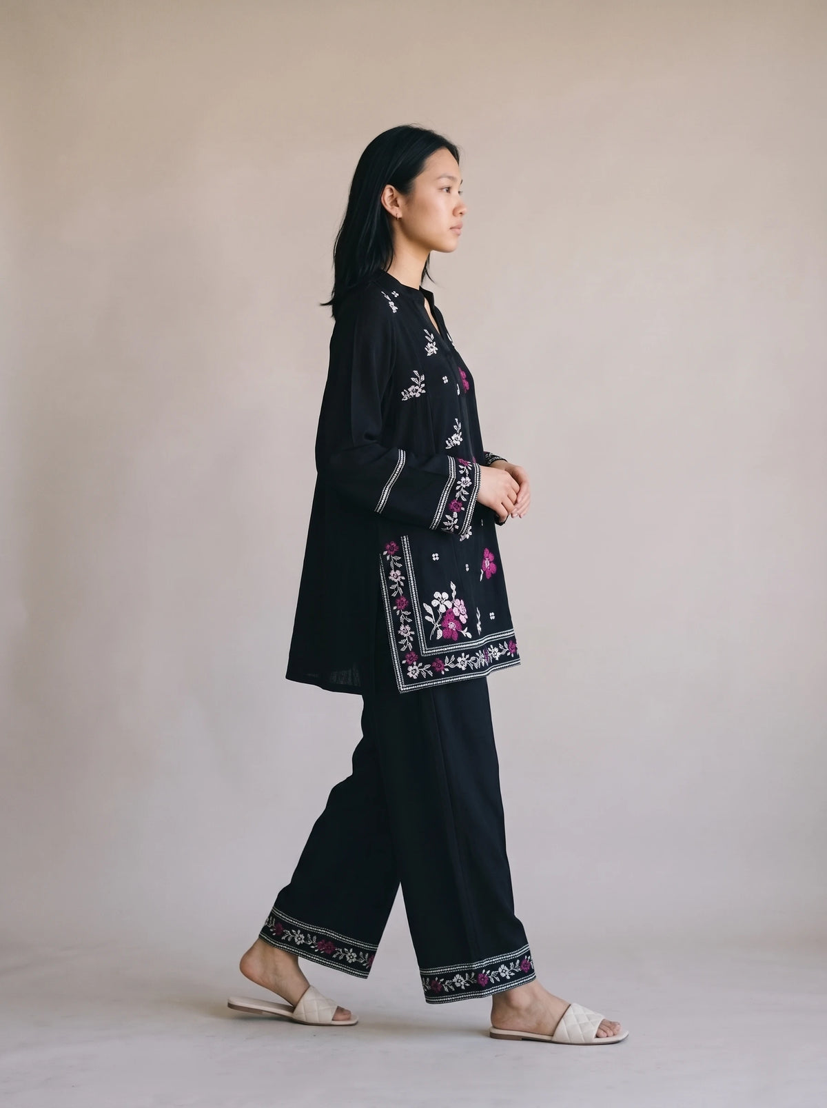 A woman in a black embroidered tunic and wide-leg pants set with floral details, walking in profile against a plain beige studio background.