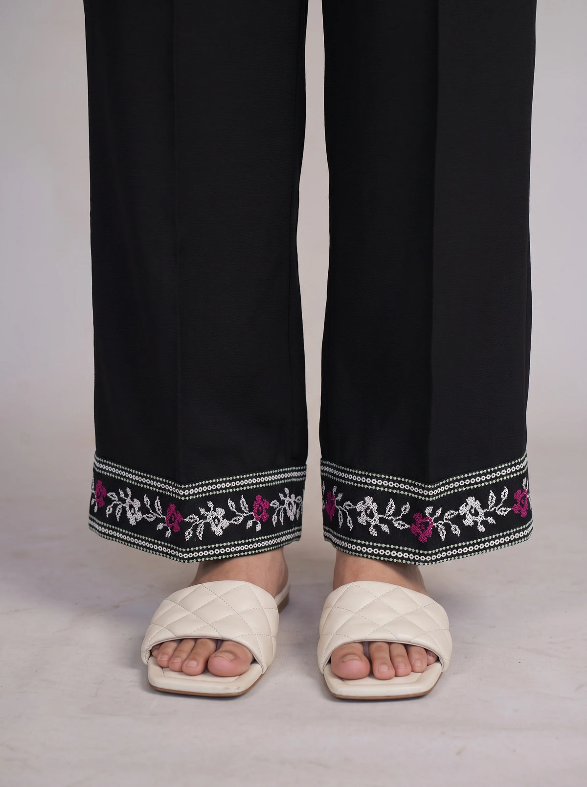 Black wide-leg trousers with a decorative embroidered hem featuring white and pink floral patterns, shown on a model wearing off-white quilted slide sandals.