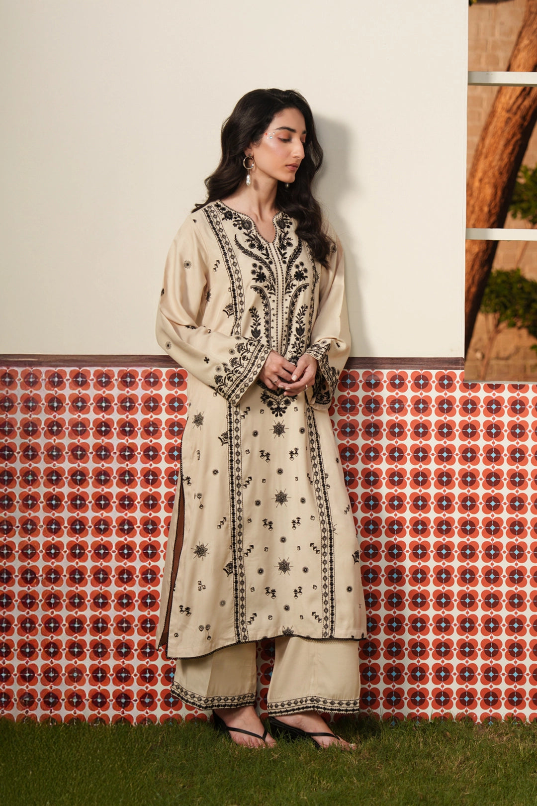 A woman wearing a beige embroidered Pakistani 2 piece suit with intricate black threadwork on the kurta and a matching trouser, standing against a red geometric backdrop — Beyond East Fall Winter 2026