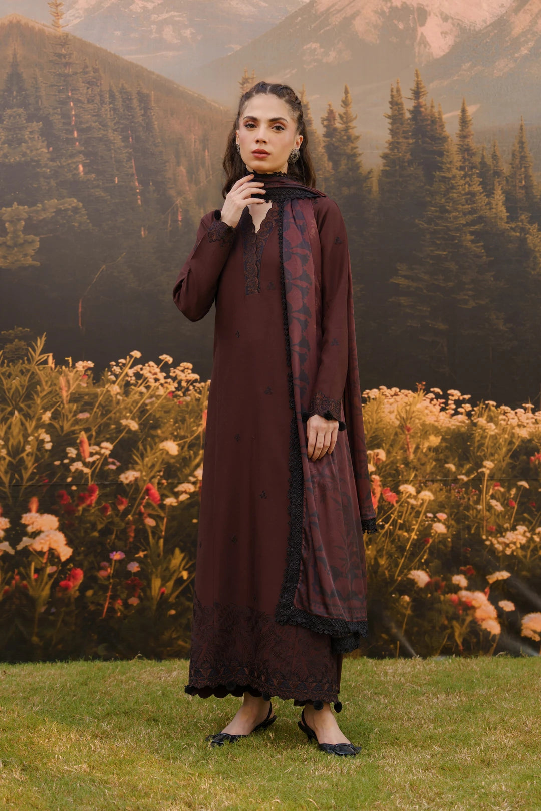 Woman wearing a dark brown embroidered linen suit with dupatta, standing in a wildflower meadow with mountains in the background — Beyond East Winter Tale collection