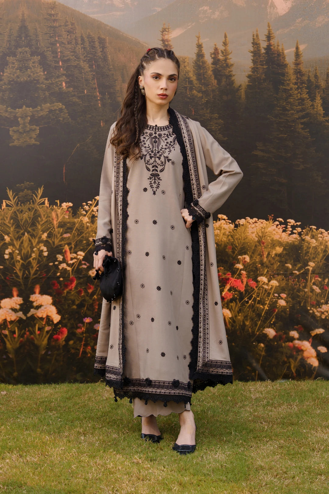 Woman wearing a grey linen suit with black floral embroidery on the front panel and sleeves, paired with a grey and black dupatta, standing in an autumn garden with mountain backdrop — Beyond East Winter Tale collection