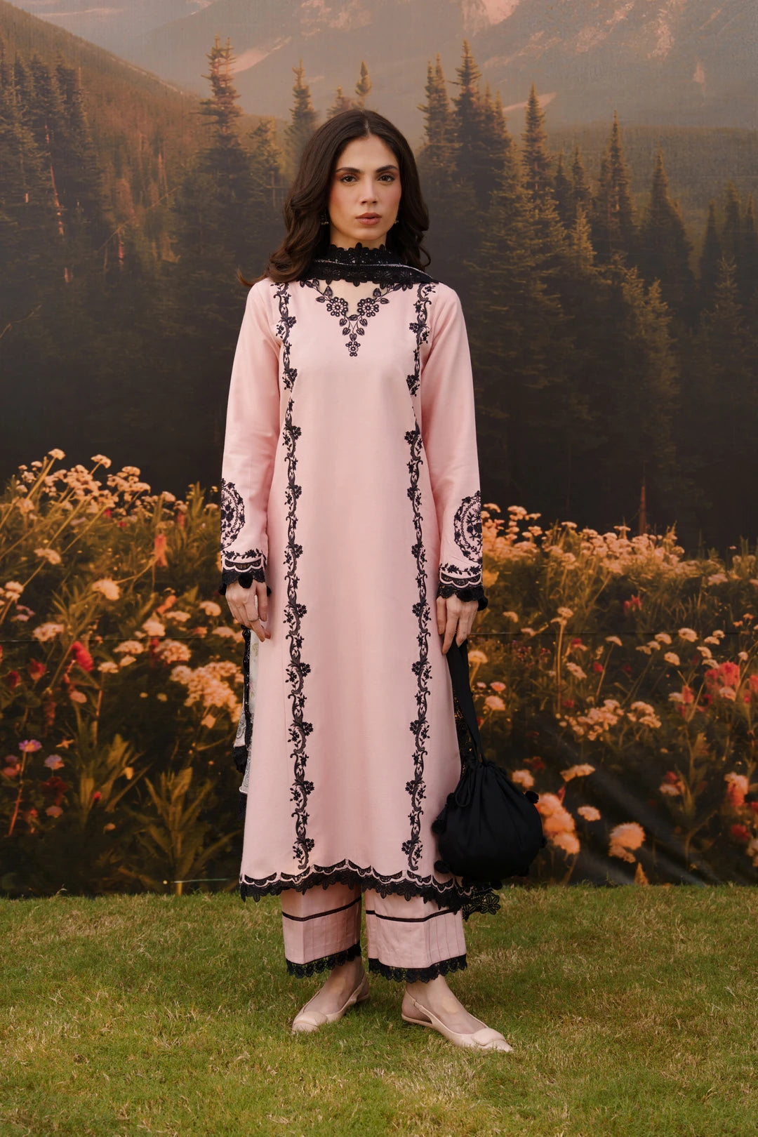 A woman stands outdoors in front of a scenic backdrop of mountains, pine trees, and flowers, wearing a light pink traditional outfit with black embroidered detailing and holding a black handbag.