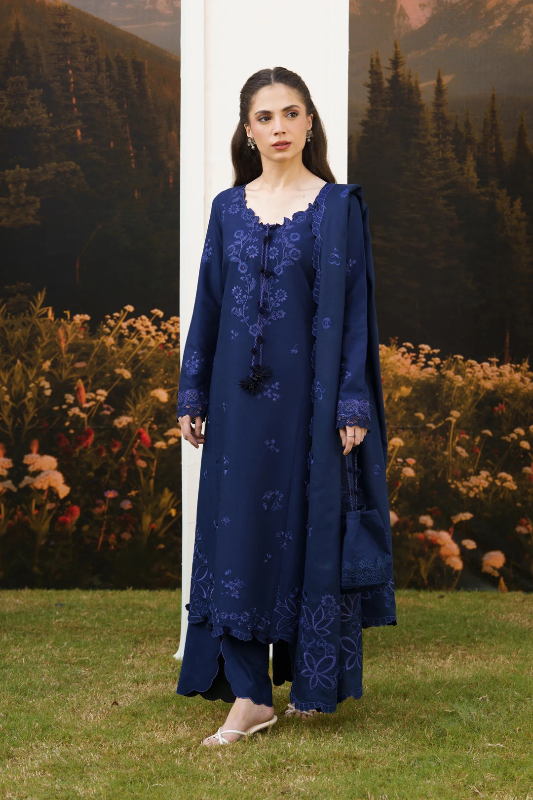 A woman modeling a navy blue traditional outfit with matching embroidered dupatta and trousers, standing outdoors in front of a scenic backdrop of trees and flowers.