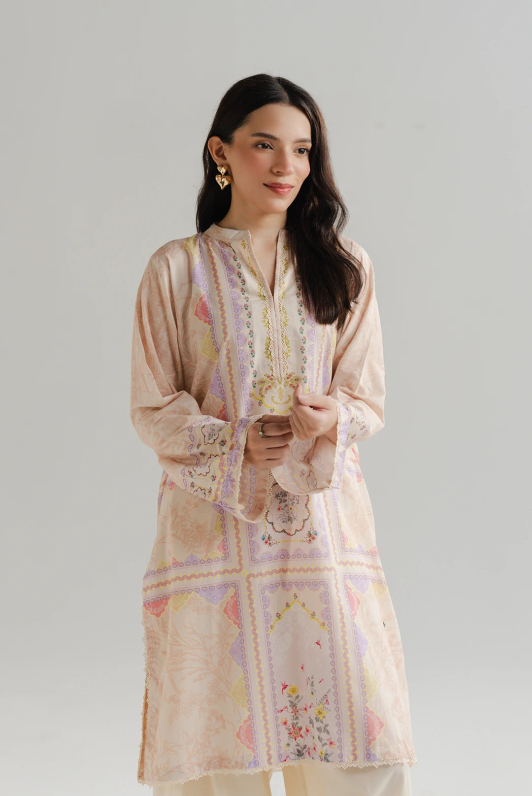 A woman wearing a pastel pink printed lawn kurti with floral and geometric embroidery, paired with white straight trousers and gold earrings — Beyond East Pakistani lawn collection