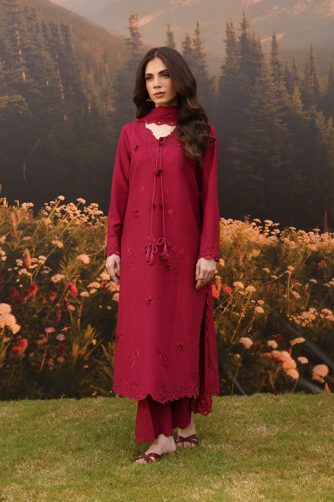 Woman wearing a deep red embroidered linen suit with flowing silhouette, standing in a field of wildflowers with mountains in the background