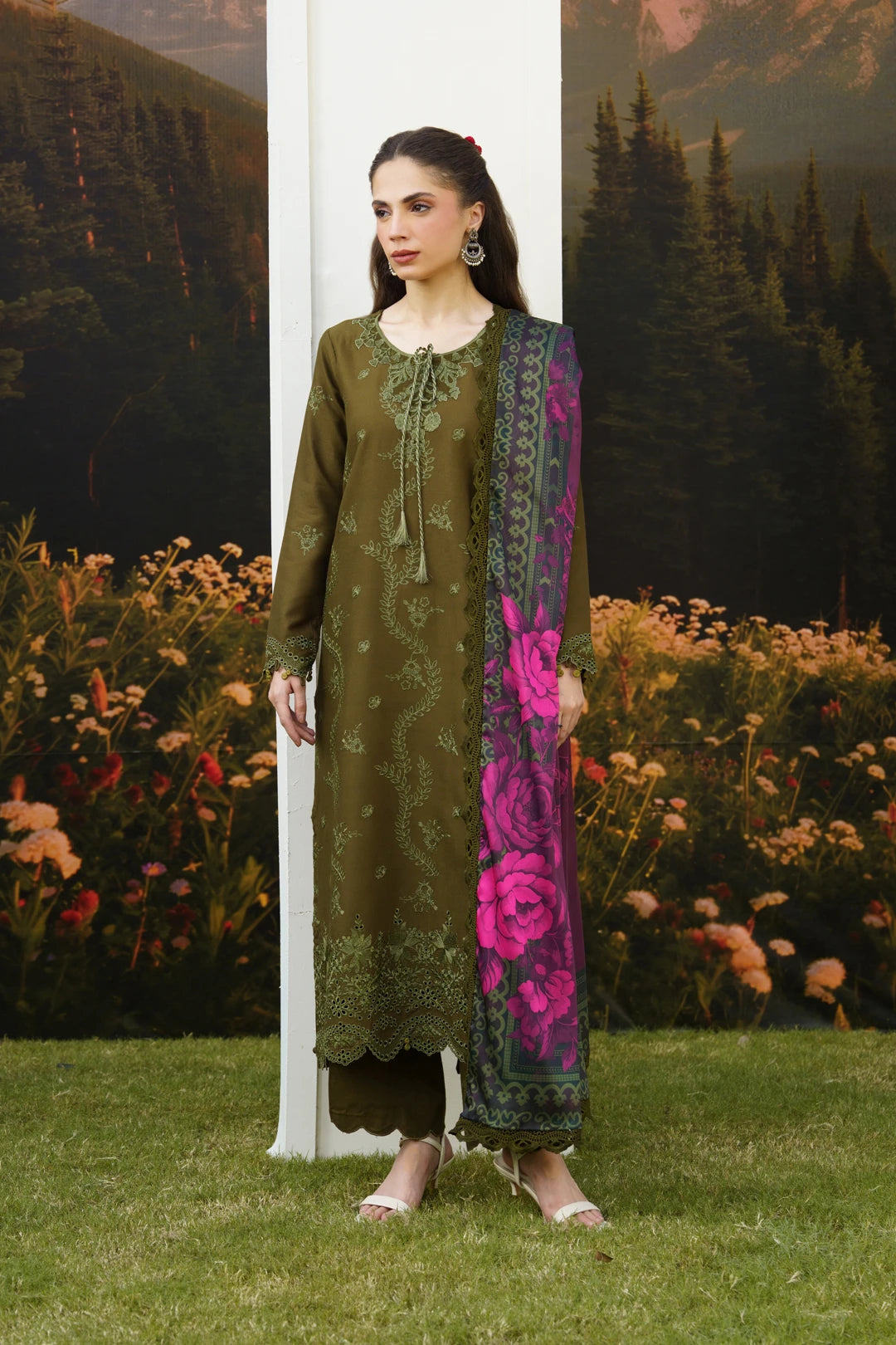 A woman models a three-piece olive green ethnic suit with floral embroidery, matching trousers, and a vibrant magenta and green floral printed dupatta, set against a scenic backdrop of grass, flowers, and a forest.